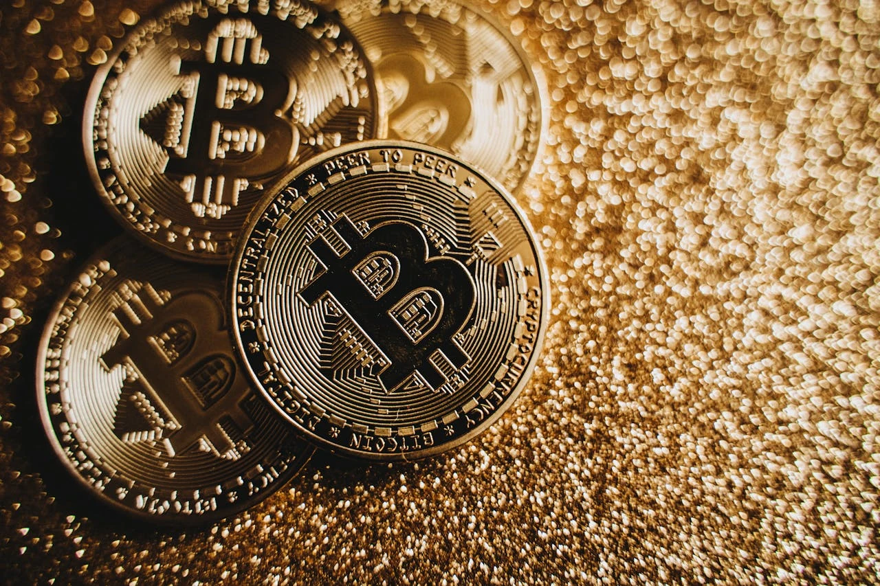 Gold-toned Bitcoin cryptocurrency coins lying on a glittering gold background, with detailed engravings visible showing the Bitcoin logo and "PEER TO PEER" text. The image has a warm sepia tone and shallow depth of field, creating a luxurious, premium aesthetic.