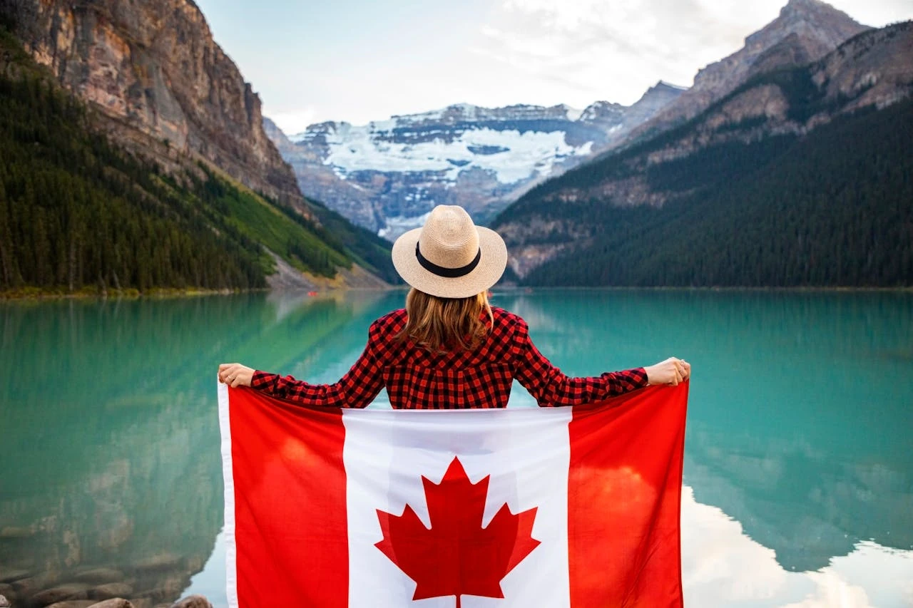 A person wearing a plaid red and black flannel shirt and straw hat holds up a Canadian flag while looking out at Lake Louise in Banff National Park. The turquoise glacial lake is surrounded by snow-capped Rocky Mountain peaks and dense evergreen forests. The image captures the iconic Canadian wilderness landscape and patriotic sentiment.