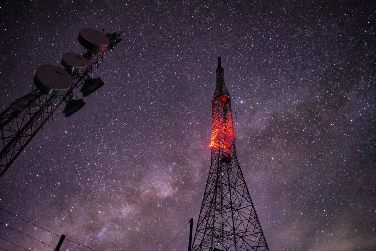 Communication towers silhouetted against a starry night sky, with one tower featuring glowing red lights along its structure while satellite dishes are visible on a separate tower to the left. The Milky Way galaxy is visible in the deep purple night sky background, creating a striking contrast between modern telecommunications technology and natural celestial beauty.