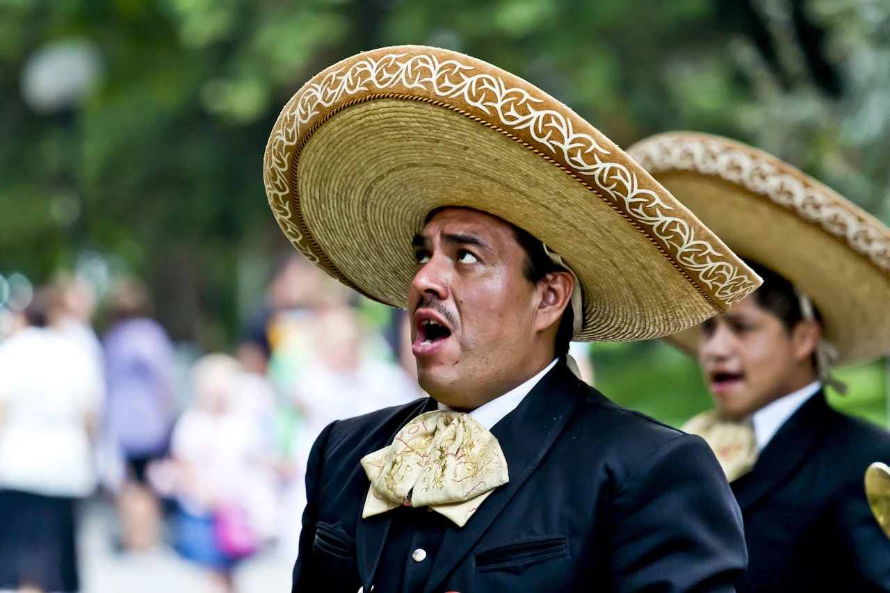 A mariachi performer wearing a traditional black charro suit and decorated sombrero singing with an open mouth during an outdoor performance