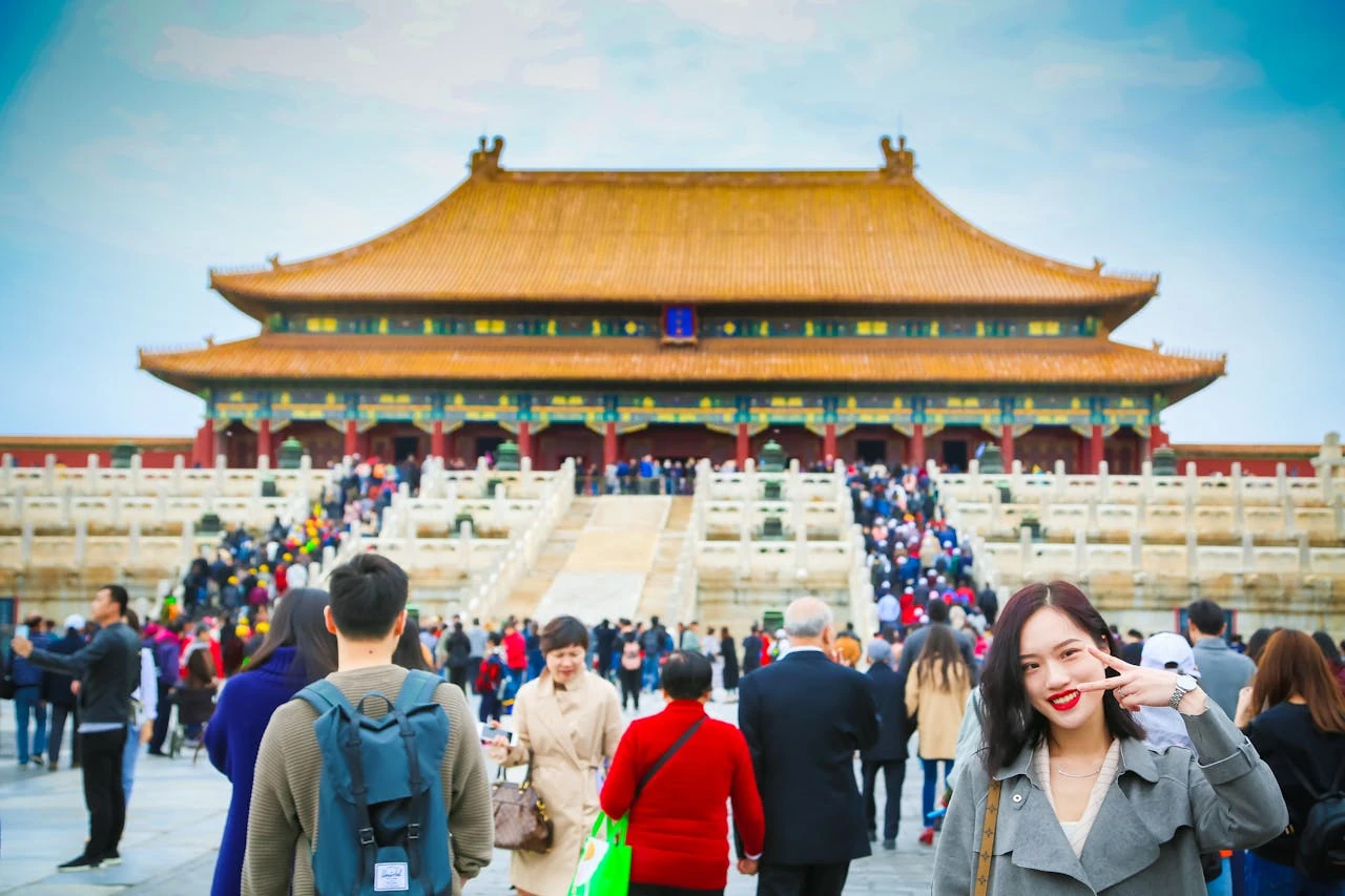 A crowded scene at the Forbidden City in Beijing, China. In the foreground, a young Asian woman makes a peace sign while smiling at the camera, wearing a grey trench coat. Behind her, numerous tourists climb the white marble stairs leading to the Hall of Supreme Harmony, a massive traditional Chinese building with a golden-tiled roof, red pillars, and ornate decorations. The sky is bright blue with light clouds.