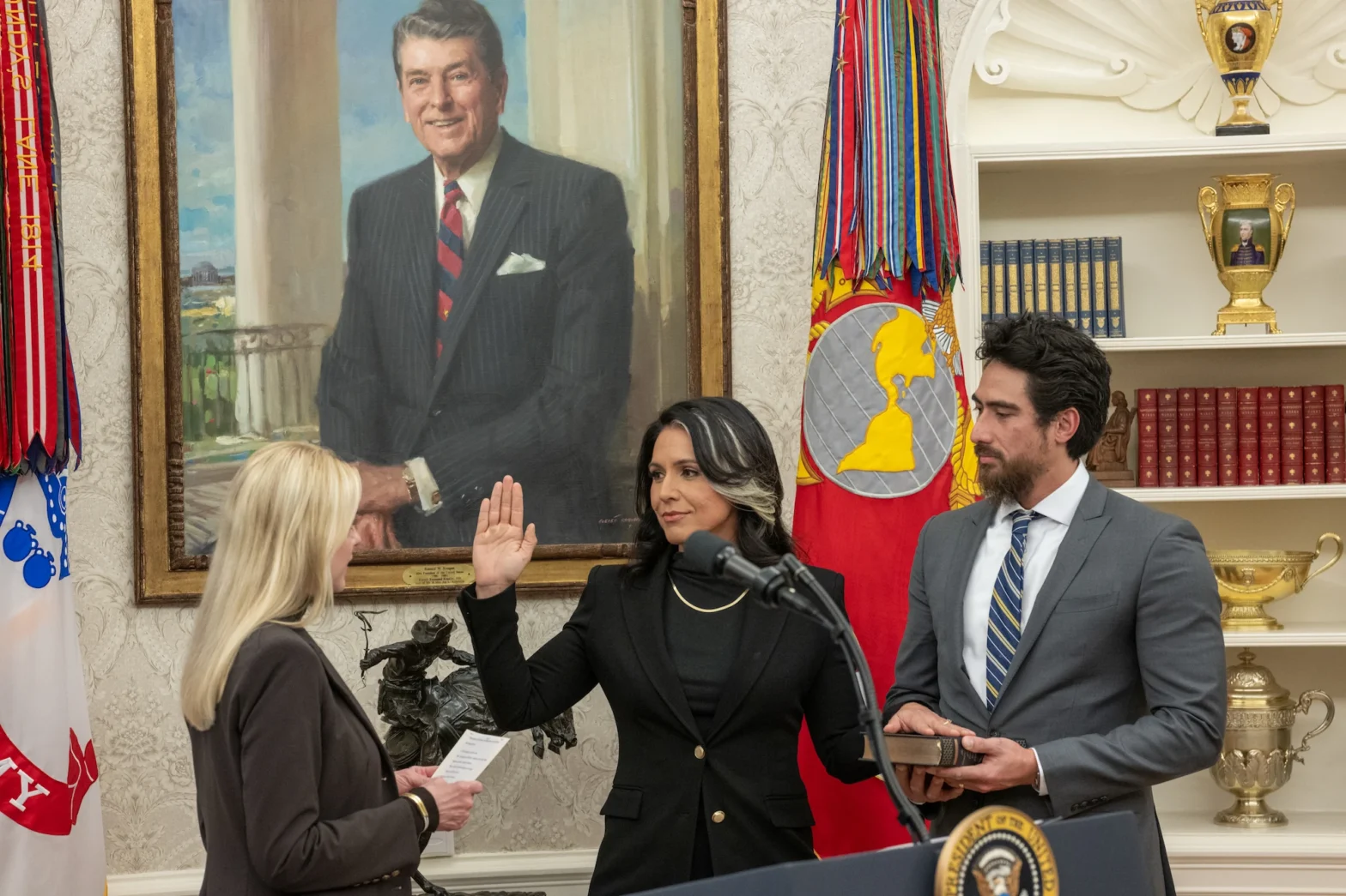 Tulsi Gabbard takes the oath of office as Director of National Intelligence in the White House, raising her right hand while being sworn in. Her husband Abraham Williams holds a Bible to her left. Attorney General Pam Bondi administers the oath. A portrait of President Ronald Reagan hangs prominently on the wall behind them, and military flags and decorative golden urns are visible in the background. The ceremony takes place in front of a white wall with ornate wallpaper patterns.