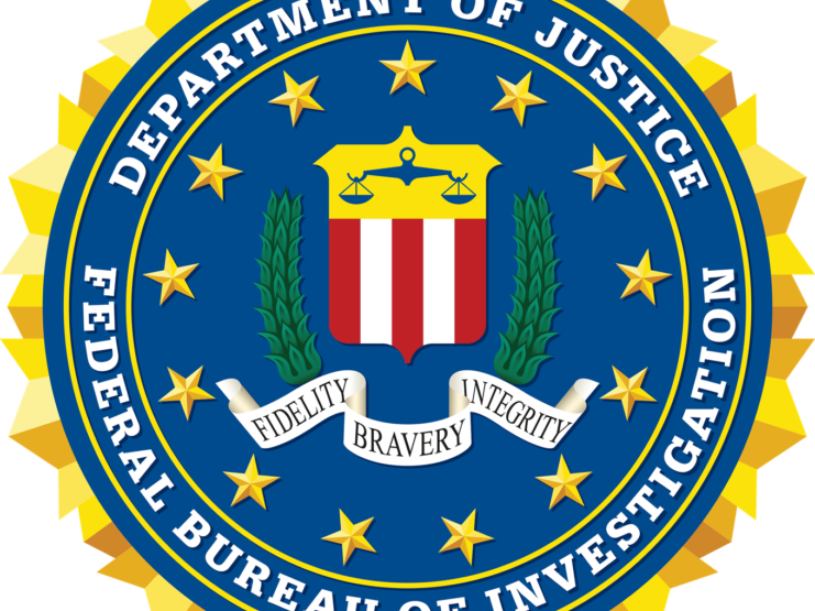 The Federal Bureau of Investigation (FBI) seal featuring a blue circular center with 13 golden stars, encircled by a golden sunburst border. The center contains a red and white striped shield topped with golden scales of justice, flanked by green laurel branches. A banner below displays the FBI's motto 