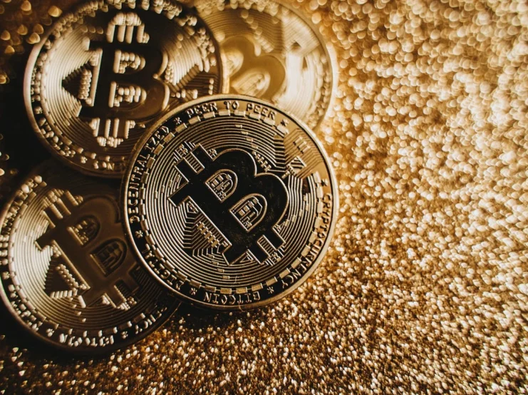 Gold-toned Bitcoin cryptocurrency coins lying on a glittering gold background, with detailed engravings visible showing the Bitcoin logo and 