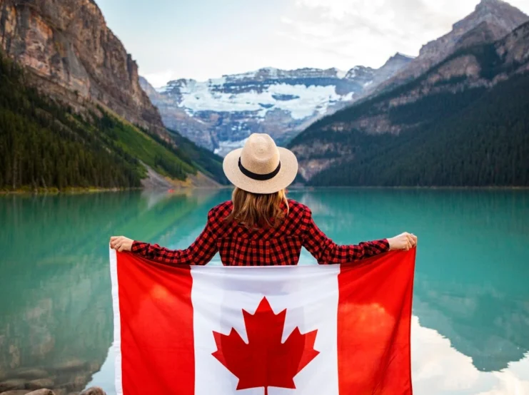 A person wearing a plaid red and black flannel shirt and straw hat holds up a Canadian flag while looking out at Lake Louise in Banff National Park. The turquoise glacial lake is surrounded by snow-capped Rocky Mountain peaks and dense evergreen forests. The image captures the iconic Canadian wilderness landscape and patriotic sentiment.