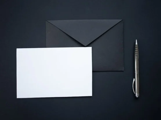 Minimalist composition on a black background featuring a black envelope, blank white paper or card, and a silver metallic pen arranged in a clean, modern layout. The items are photographed from above against a dark surface, creating a stark contrast between light and dark elements.