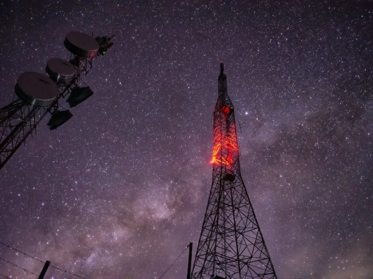 Communication towers silhouetted against a starry night sky, with one tower featuring glowing red lights along its structure while satellite dishes are visible on a separate tower to the left. The Milky Way galaxy is visible in the deep purple night sky background, creating a striking contrast between modern telecommunications technology and natural celestial beauty.