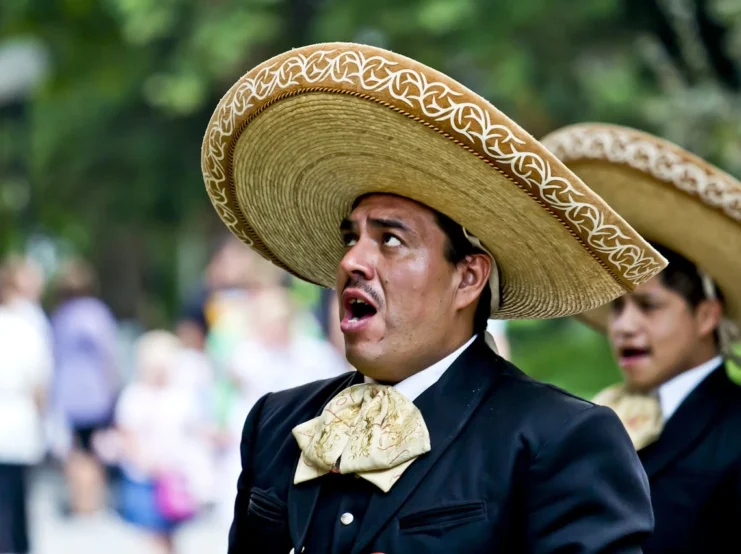 A mariachi performer wearing a traditional black charro suit and decorated sombrero singing with an open mouth during an outdoor performance