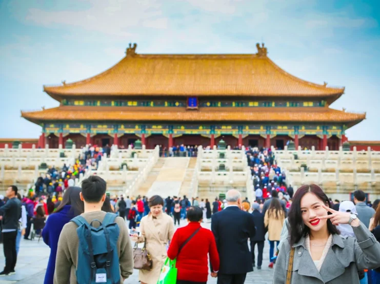 A crowded scene at the Forbidden City in Beijing, China. In the foreground, a young Asian woman makes a peace sign while smiling at the camera, wearing a grey trench coat. Behind her, numerous tourists climb the white marble stairs leading to the Hall of Supreme Harmony, a massive traditional Chinese building with a golden-tiled roof, red pillars, and ornate decorations. The sky is bright blue with light clouds.