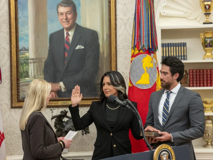 Tulsi Gabbard takes the oath of office as Director of National Intelligence in the White House, raising her right hand while being sworn in. Her husband Abraham Williams holds a Bible to her left. Attorney General Pam Bondi administers the oath. A portrait of President Ronald Reagan hangs prominently on the wall behind them, and military flags and decorative golden urns are visible in the background. The ceremony takes place in front of a white wall with ornate wallpaper patterns.